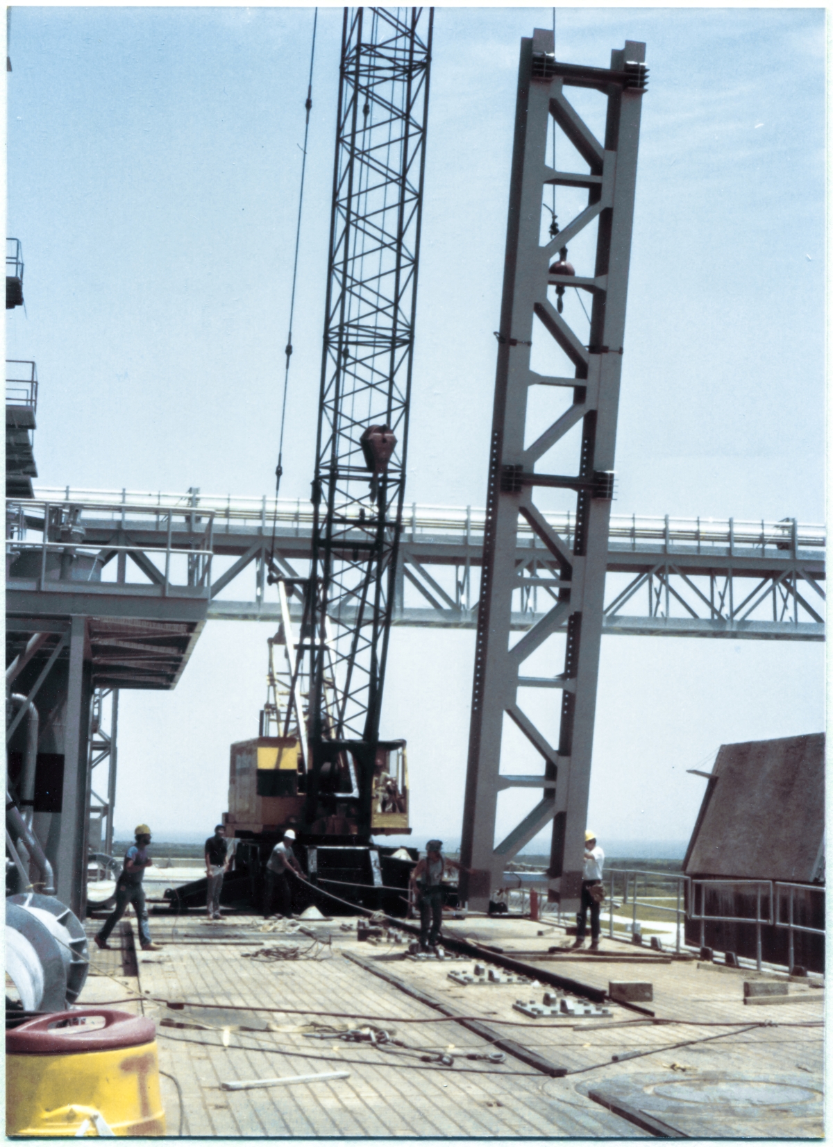 Image 076. The GOX Arm Strongback lift is now fully under way, with the strongback clear of the ground which was supporting it, now hanging in suspension, supported solely by its outré lifting sling which is attached via a set of shackles up above the headache ball and the crane's lifting hook beneath that. In this image, the headache ball can clearly be seen hanging off-center, away from the crane's jib line, its own attach hardware pulled to the side at a significant angle, bearing no load above and beyond its own weight. From this angle, the choker slings wrapped around the twin W24x104's which constitute the main body of the Strongback can be seen resting on pads at each corner of the wide-flanges to keep from chafing, possibly damaging not only the Strongback, but also the wire ropes of the slings. Photo by James MacLaren.
